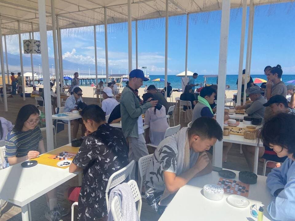 People playing Go at white tables under a canopy by a beautiful beach with mountains in the background.