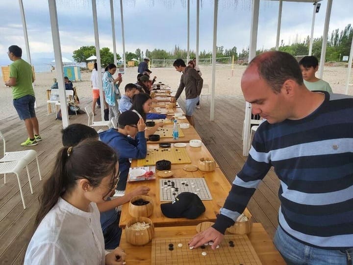 A group of young and adult players focused on Go games at wooden tables near a beach.