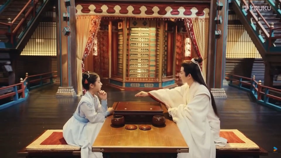 A scene from Dashing Youth showing two characters seated across a Go board in an ornate interior hall.