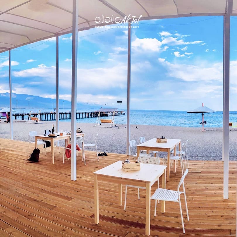 Empty white tables set up for Go games on a wooden deck overlooking the sea and pier.