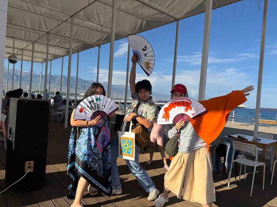 Three festival participants posing with decorated fans on a sunny terrace by Lake Issyk-Kul.