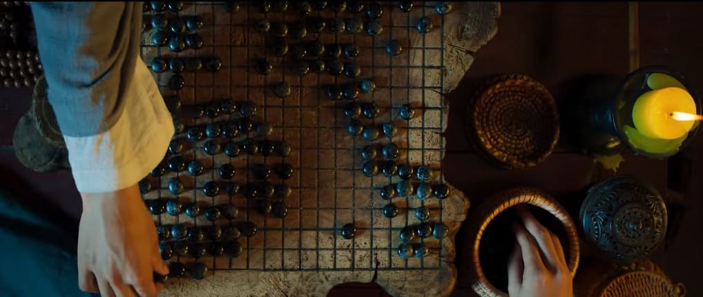 Cinematic overhead shot of a worn go board with dark stones, players’ hands, bowls on the table, and a lit candle nearby.
