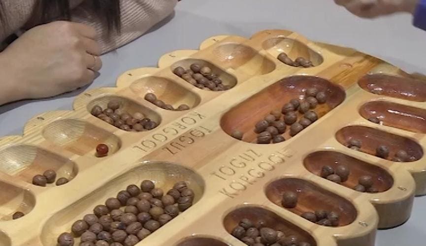 A wooden Toguz Korgool board with brown playing stones during a match.