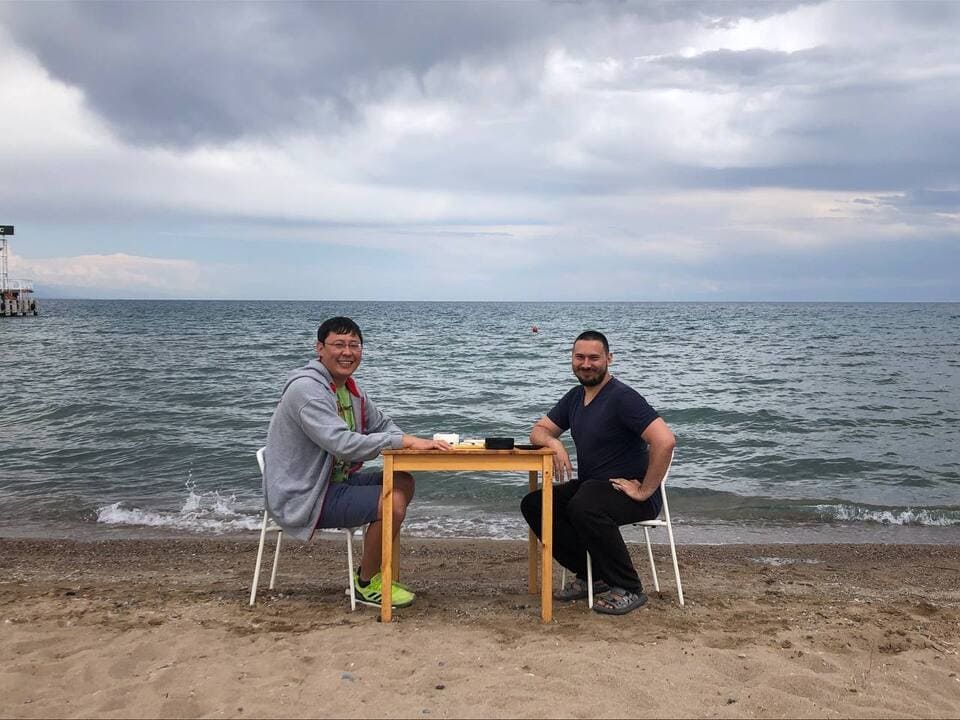 Two men playing Go at a small table on the beach with the sea in the background.
