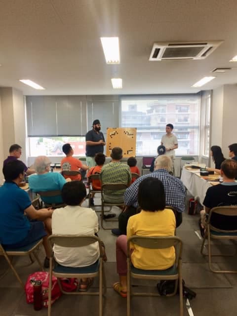 Participants sitting in a classroom while two speakers present a Go lesson with a demonstration board