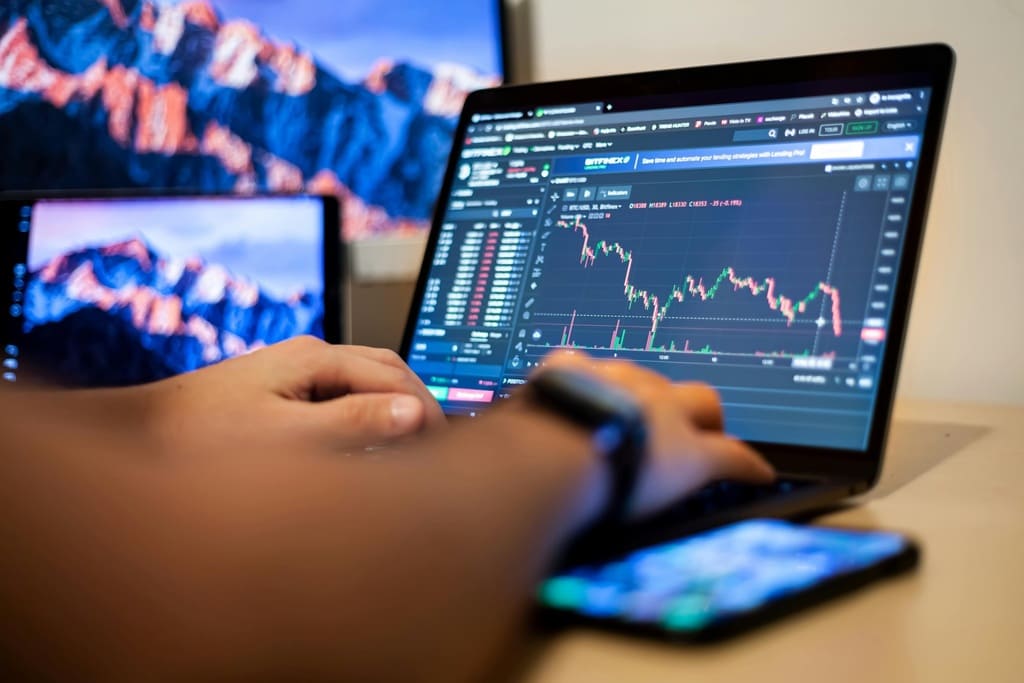 A close-up photo of a person using a laptop with stock trading charts, with a smartphone and extra screens in the background.