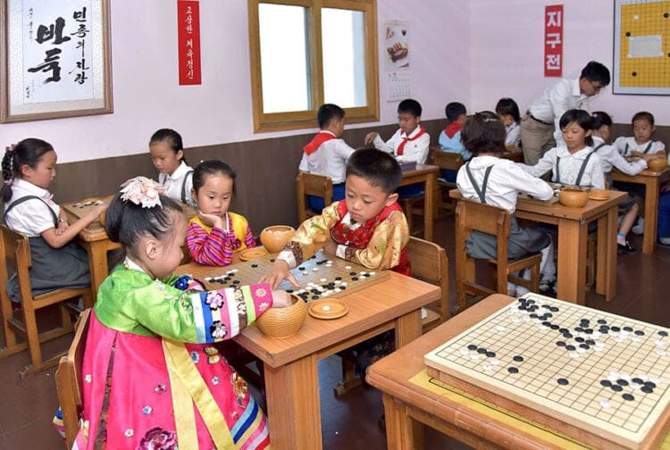 A classroom scene with children seated at wooden desks playing Go, including two children in colorful traditional clothing in the foreground.