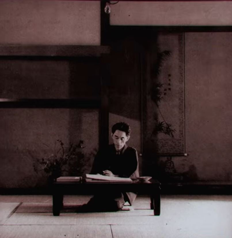 A still showing a man seated at a low table in a traditional Japanese room, writing quietly by himself.