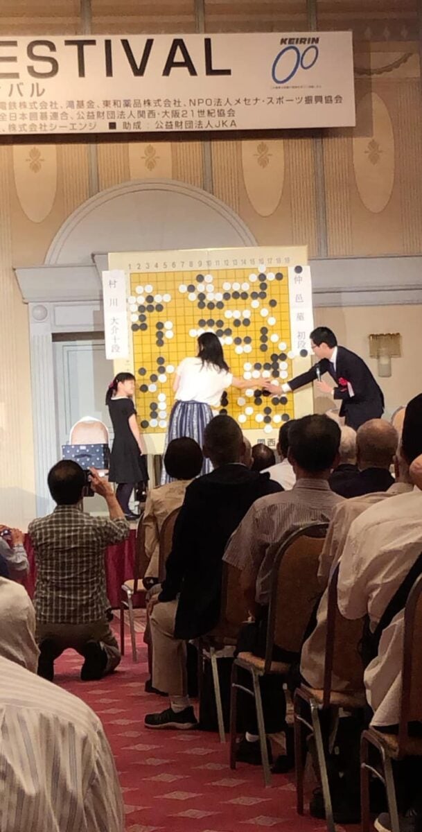 Public Go event with a large demonstration board on stage and an audience watching