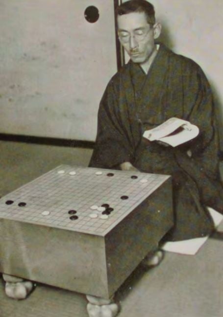 Black-and-white archival photo of a man in kimono seated beside a Go board with stones already in play.