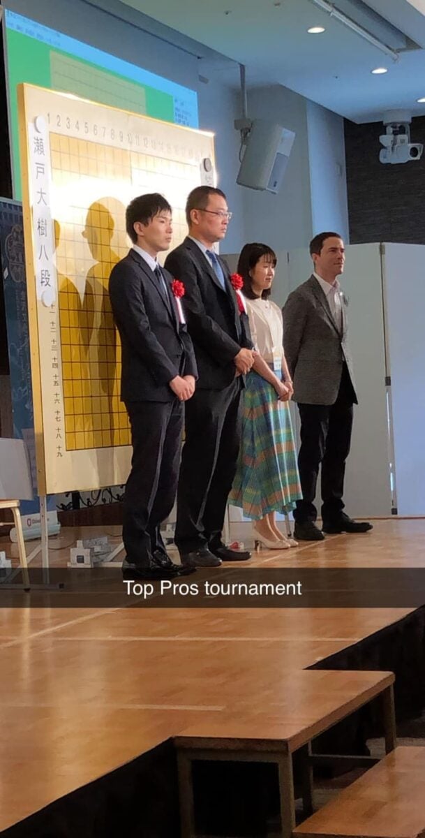 Professional Go tournament presentation with players and organizers standing beside a large board on stage