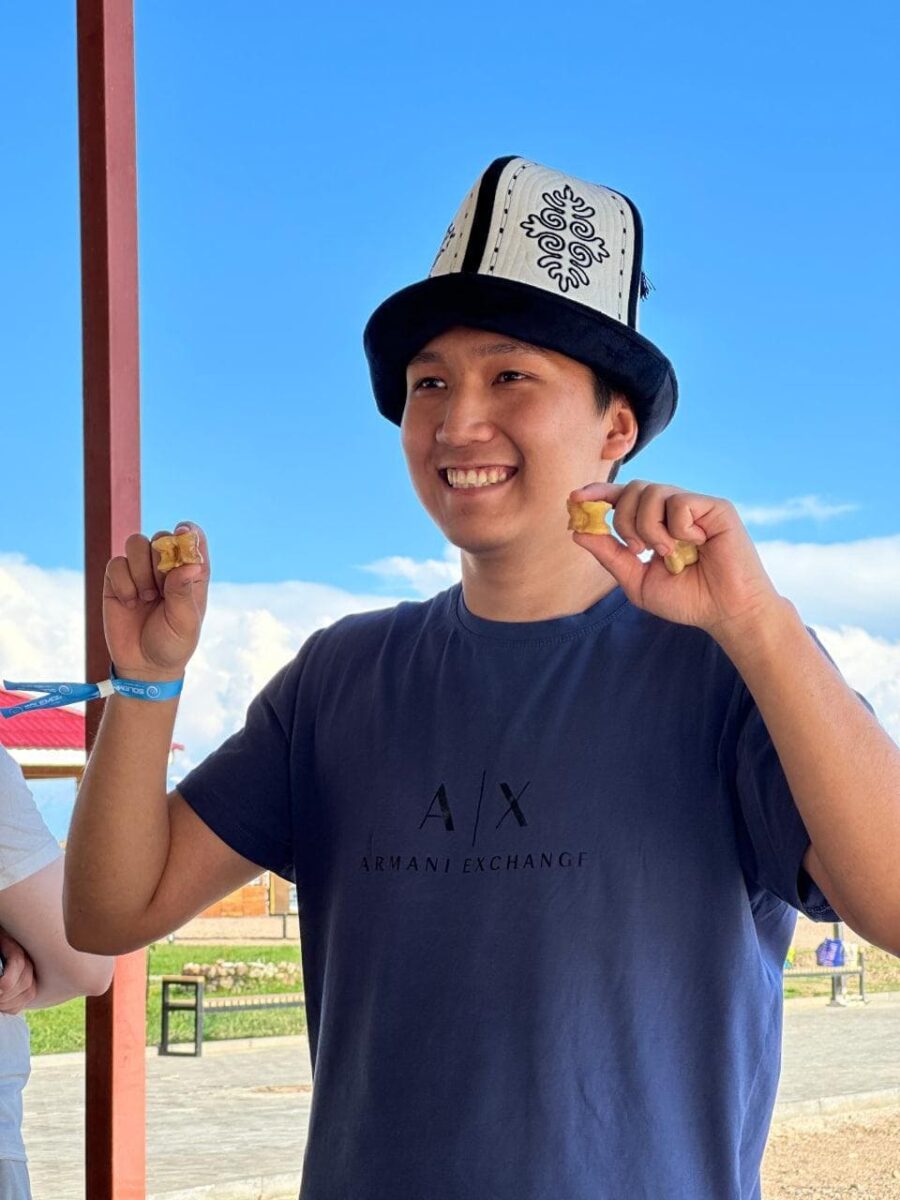 A smiling young man in a traditional Kyrgyz hat holding tokens, celebrating Go success.