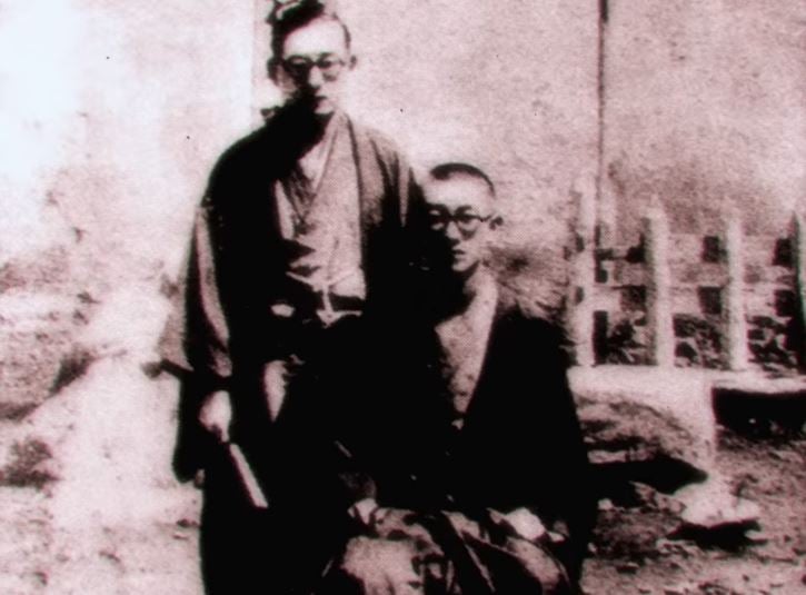 Vintage black-and-white photo of two men in traditional Japanese dress, one standing and one seated outdoors.