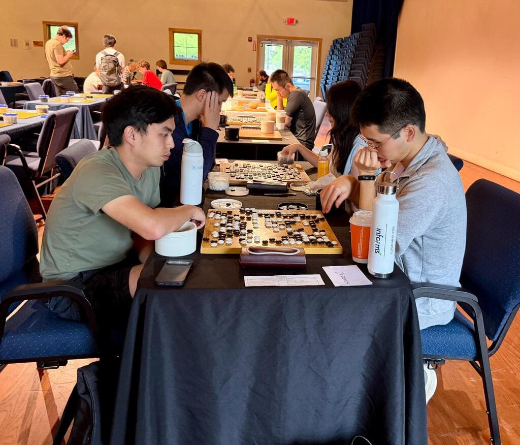 Two players studying a Go position across a tournament table, with other games in progress behind them.