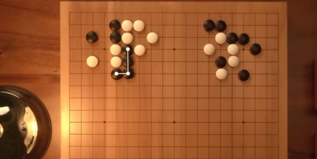 Close-up top-down view of a Go board showing clusters of black and white stones forming in two upper corners.