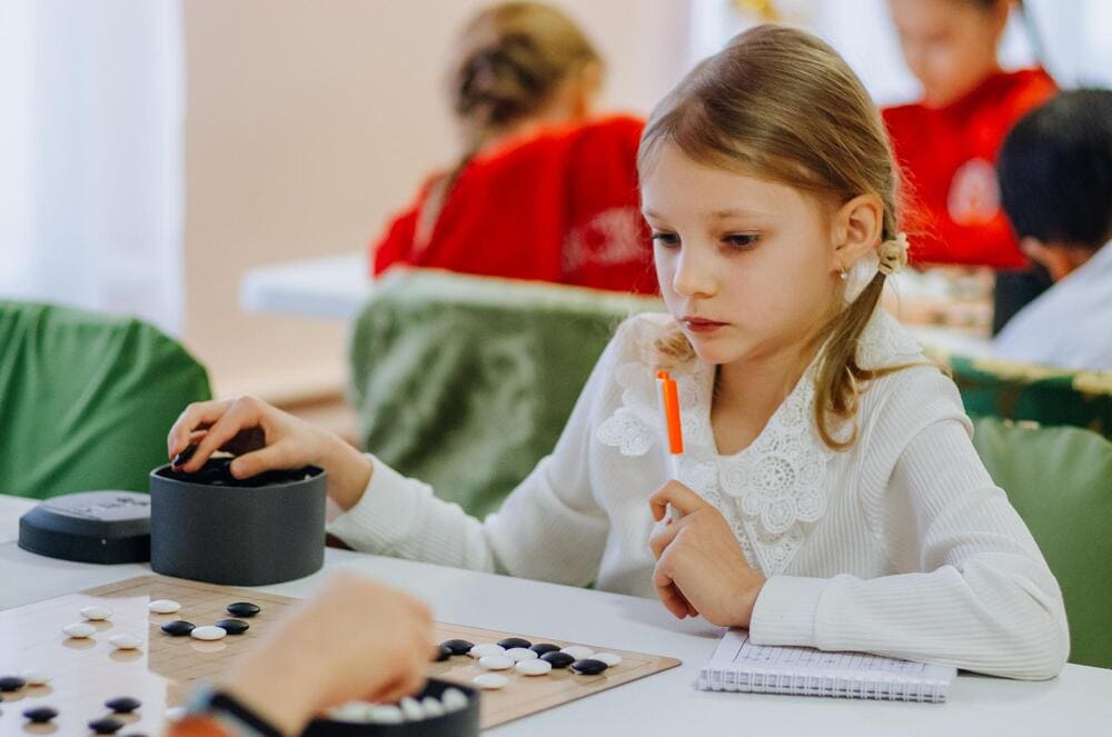 A young girl in a white dress focuses on her Go game during a children's tournament, holding a pen and looking at the board.