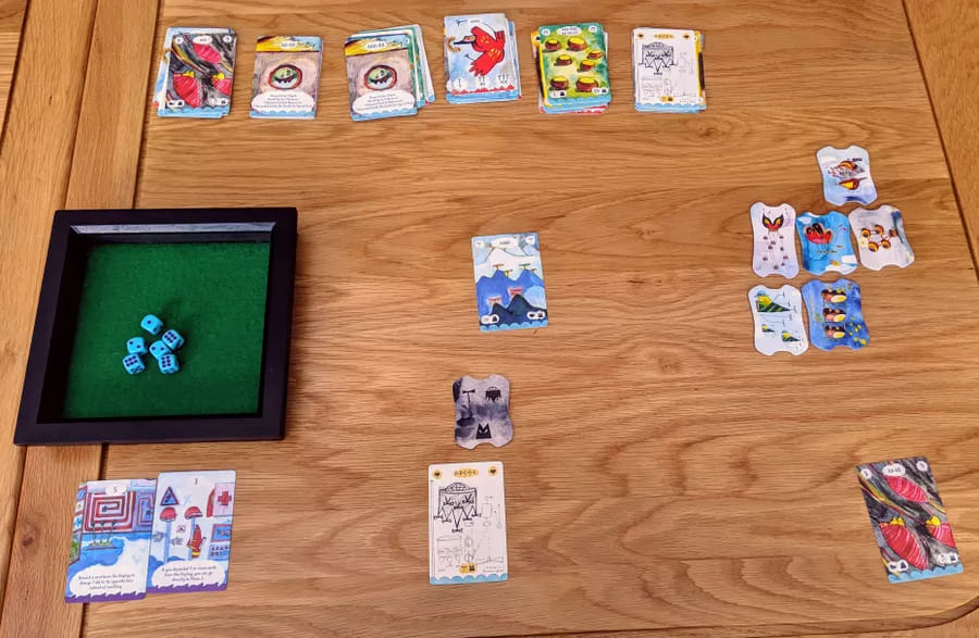 A setup of the board game Aerion, featuring dice, cards, and colorful illustrations spread out on a wooden table.