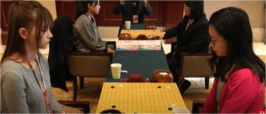 Joanne Missingham sitting across from an opponent during a Go match, focused on the board, with two other players visible in the background.