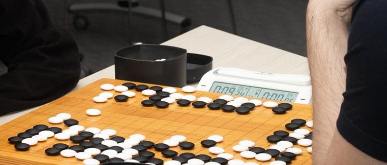 Two players intensely focused on a Go game with a digital timer displaying remaining time and scattered black and white stones on the wooden Go board.