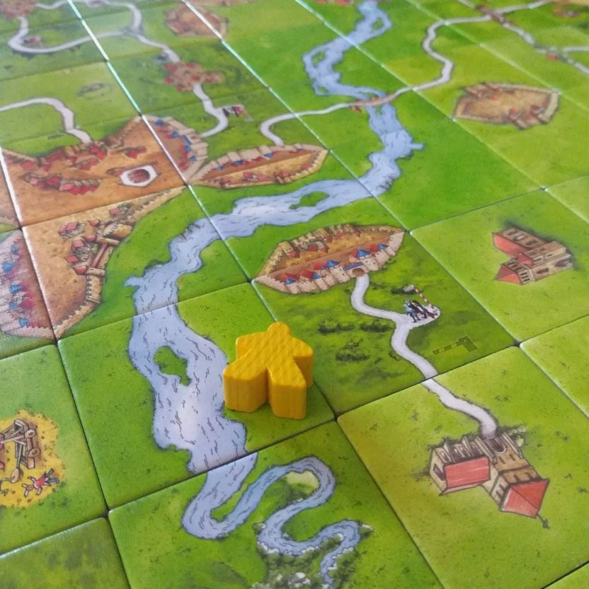 A detailed view of a Carcassonne board game with tiles depicting landscapes, rivers, and a yellow meeple placed on the board.