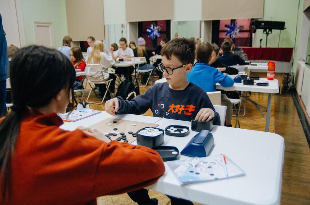 A young boy wearing glasses plays Go against an opponent in a classroom setting, surrounded by other children engaged in matches.