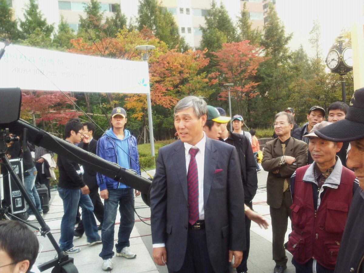 Cho Hun-Hyun, a renowned professional Go player, surrounded by fans and participants at a public Baduk event. The vibrant autumn foliage and event banners add to the lively atmosphere.