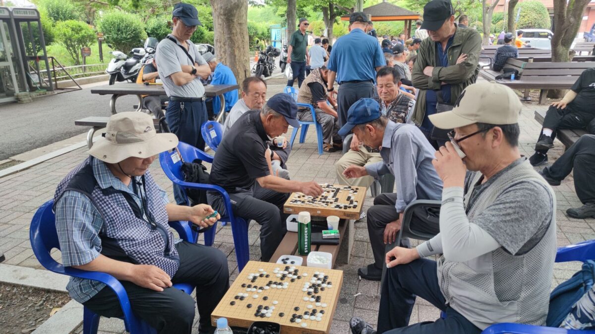 A group of elderly men deeply engaged in playing Go in an outdoor park setting. The relaxed environment reflects the cultural tradition of Baduk as a social and intellectual pastime.