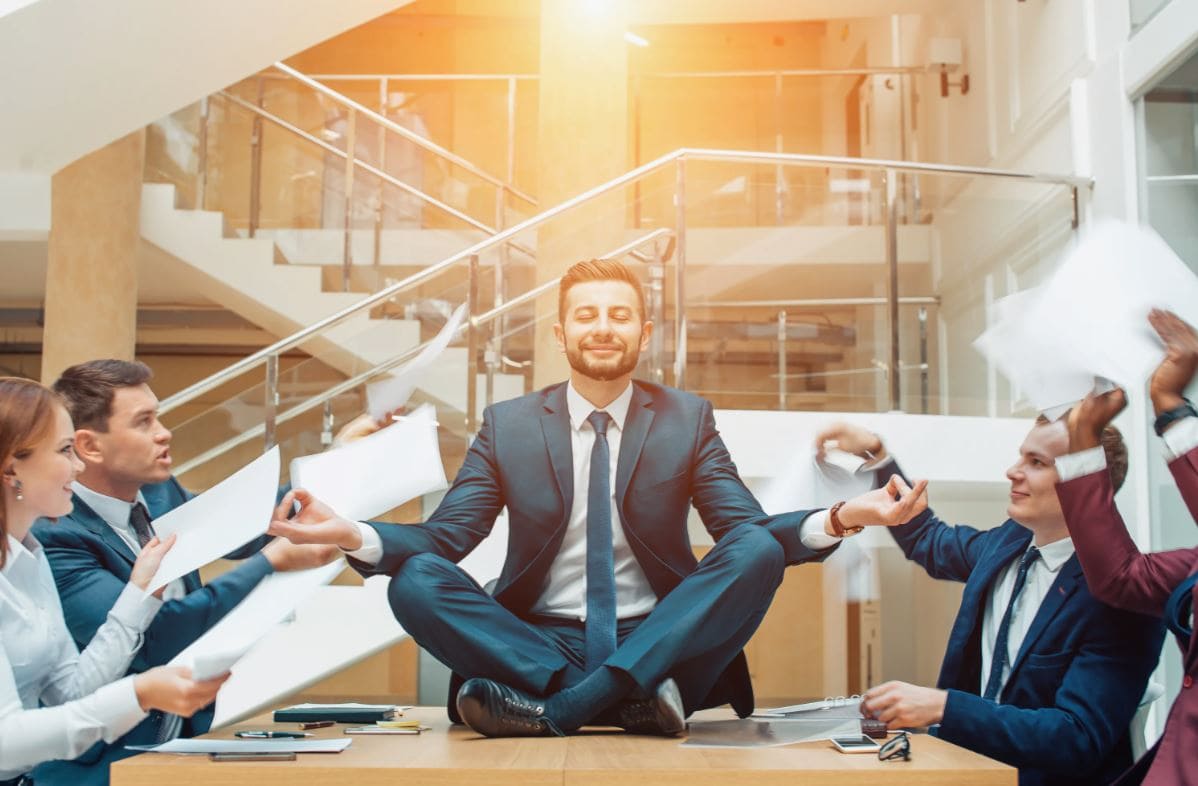 A man meditating calmly on a desk in a busy office while colleagues around him are in chaos, representing stress management.