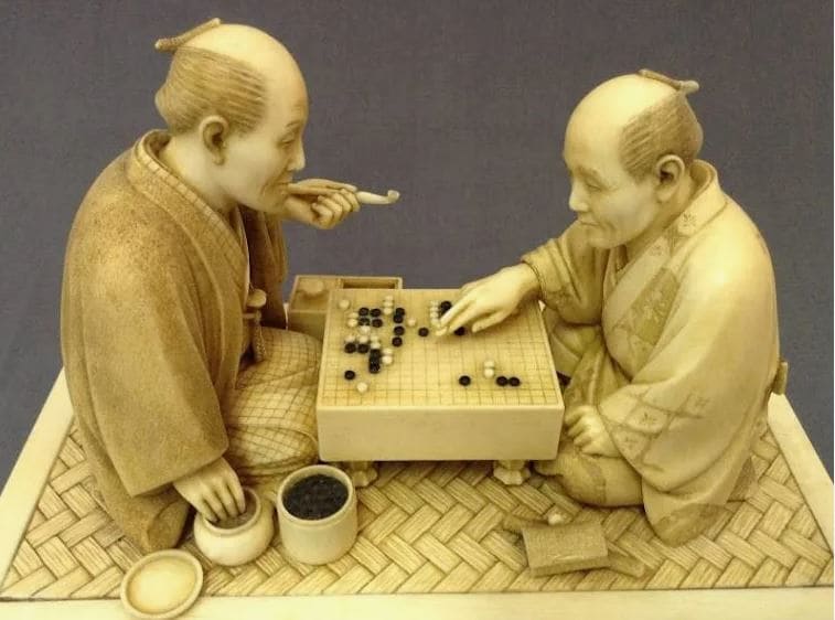 A detailed sculpture of two traditional Japanese men playing a game of Go, one of them holding a pipe, representing a classic scene of the game's cultural heritage.