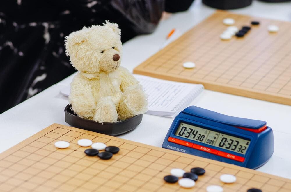 A plush teddy bear sits inside a Go bowl next to a game board and a digital clock, adding charm to the tournament setting.