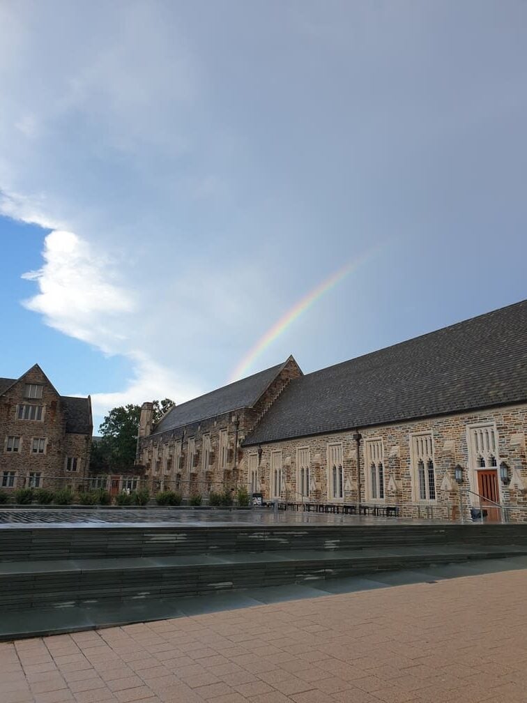 A tall building photographed from below, with a rainbow visible in the cloudy sky.