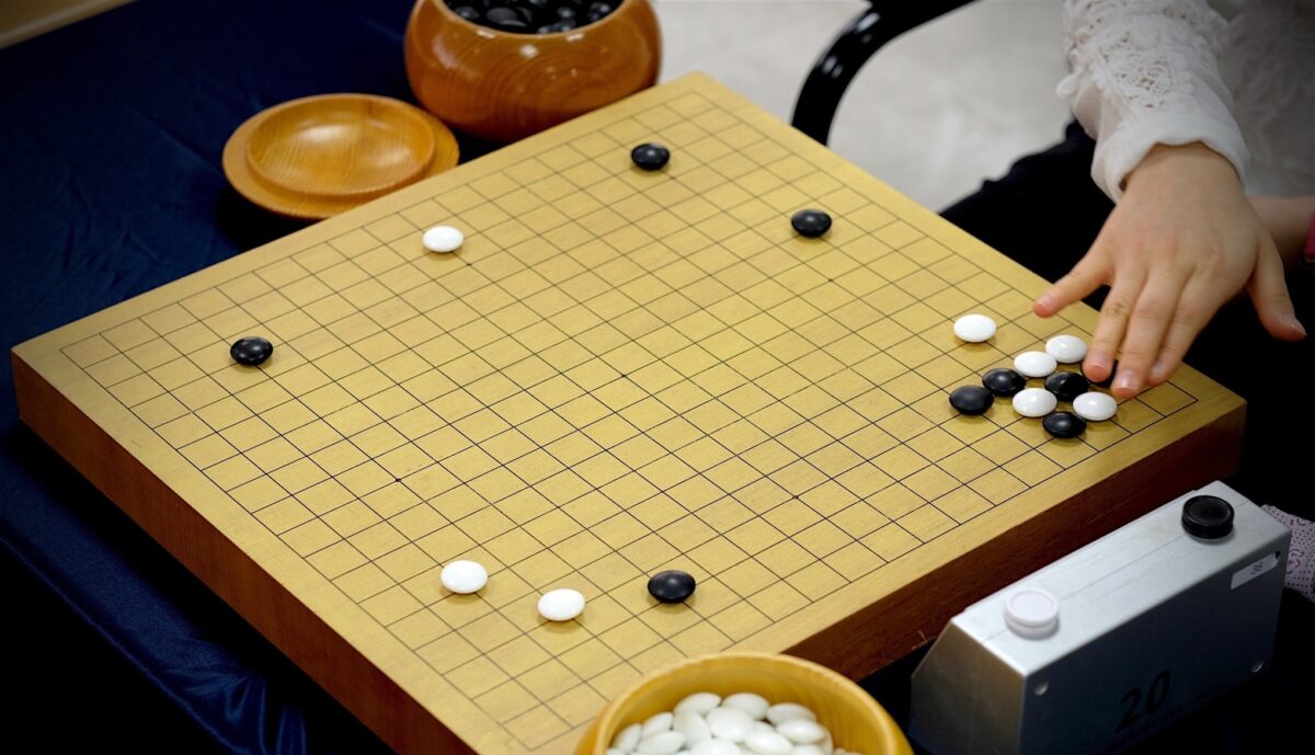 A close-up of a Go board featuring a strategic opening setup with black and white stones, wooden bowls of stones, and a digital timer.