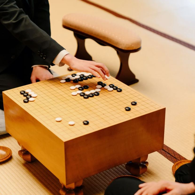 Two people playing Go on a traditional wooden board with black and white stones.