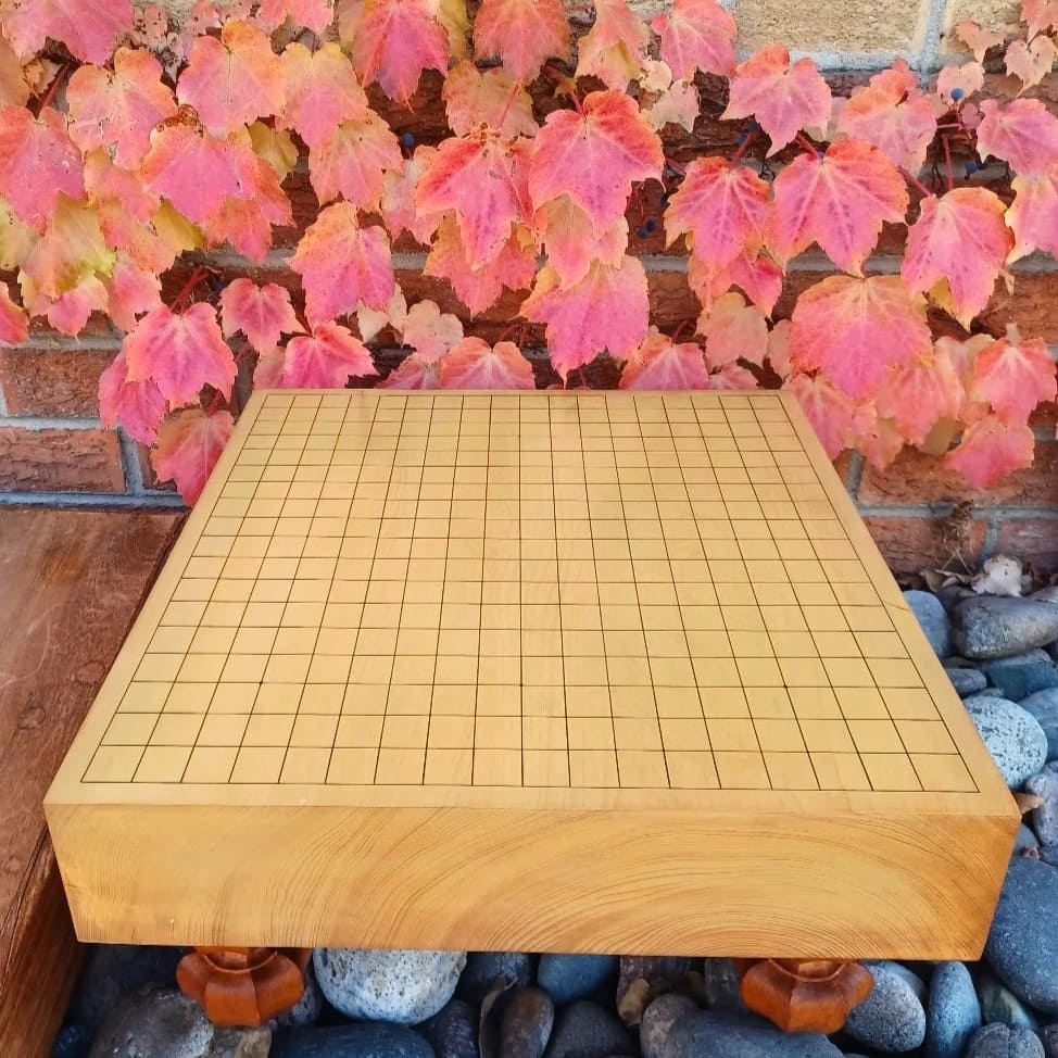 A traditional kaya Go board displayed on rocks with colorful autumn leaves in the background