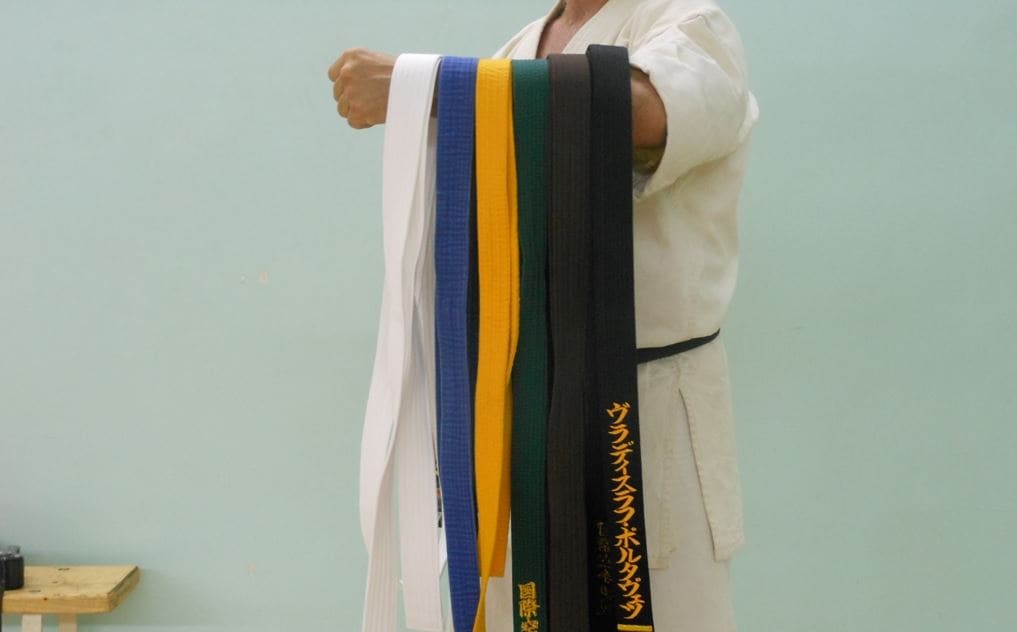 A martial artist holding multiple colored belts, including white, yellow, blue, green, and black, in a training environment.