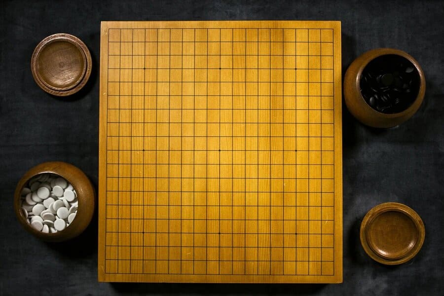 A top-down view of a traditional Go board (goban) with two wooden bowls filled with black and white stones.