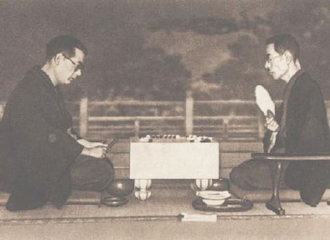 A historic black-and-white photo of Honinbo Shusai and Kitani Minoru seated during a Go game, with traditional Japanese interior in the background.