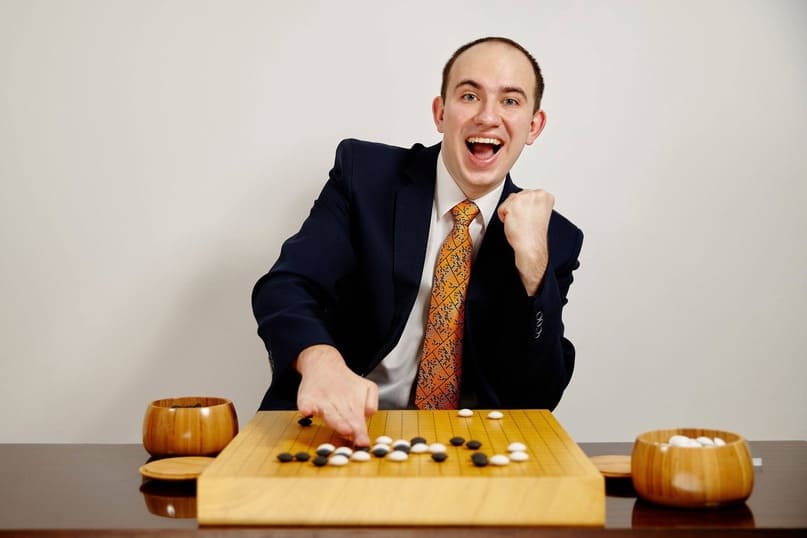 European Go professional Mateusz Surma in a suit and vibrant tie, smiling and clenching his fist in excitement while pointing to a move on the Go board, symbolizing a victorious moment.