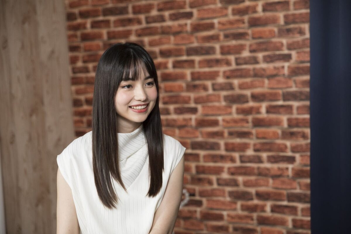 Joanne Missingham smiling in a white dress, sitting in front of a brick wall during an interview.