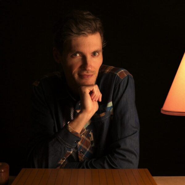A thoughtful man sits by a Go board and lamp in a dimly lit room