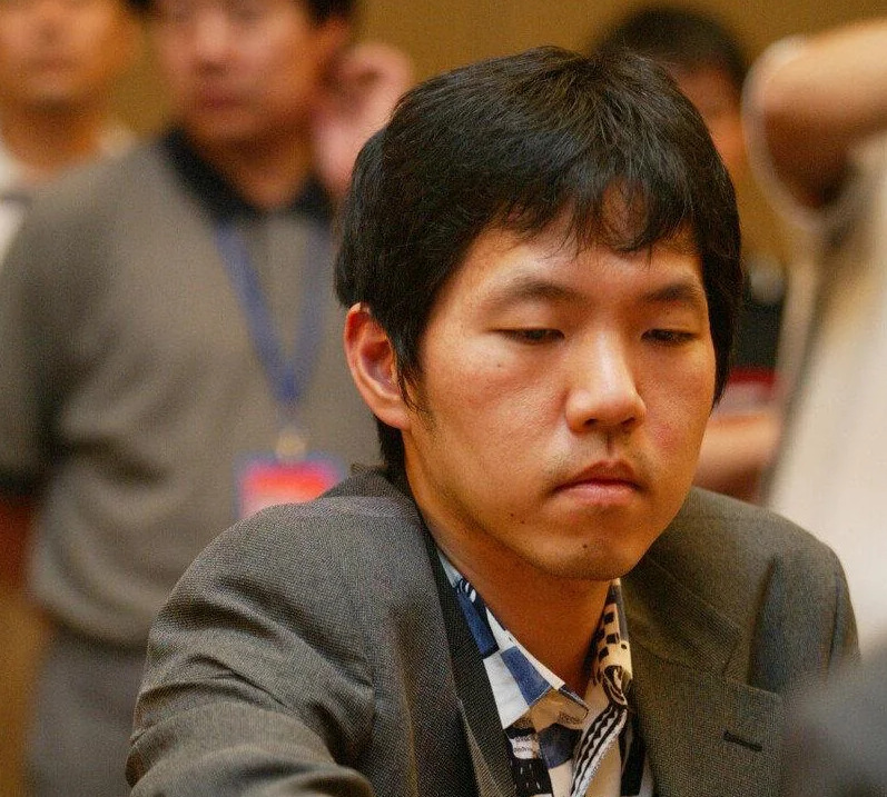 Lee Changho, the renowned "Stone Buddha" of Go, wearing a suit and focused intently as he plays a match, preparing to place a stone on the Go board.