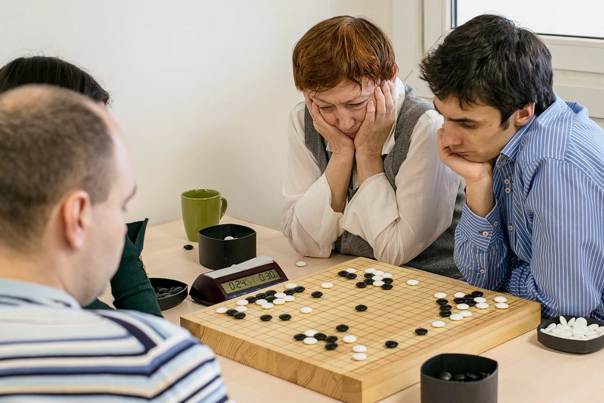 Four individuals engaged in a focused Go game with a clock in the background, capturing the intensity and strategic depth of the board game.