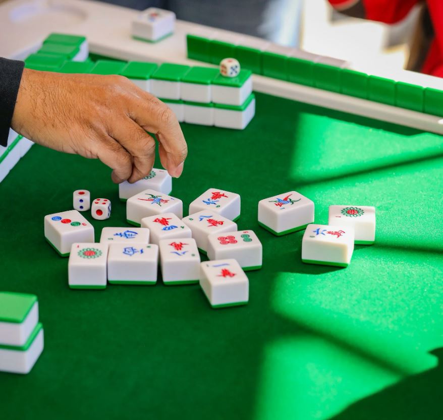 A close-up of a person’s hand playing Mahjong, showing the tiles and dice on a green table.