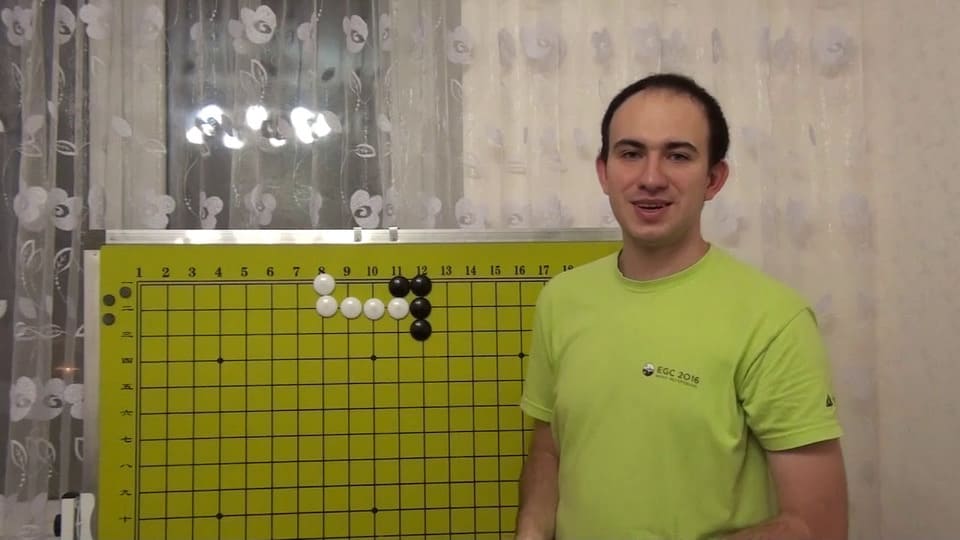 Mateusz Surma standing beside a large yellow Go board, explaining a joseki sequence during a Go lesson or workshop.