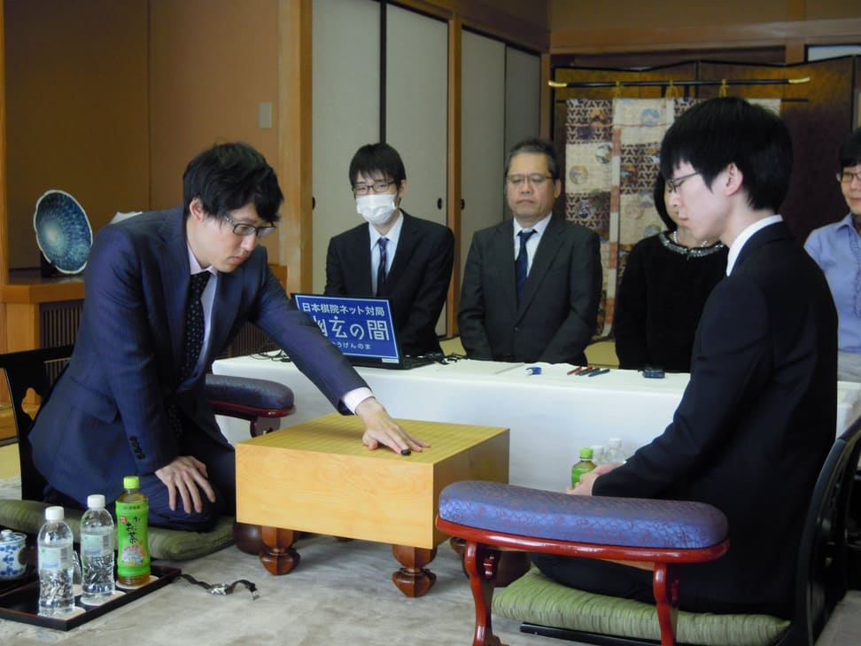 A formal Go match between two professional players in suits, observed by a panel of judges and officials.