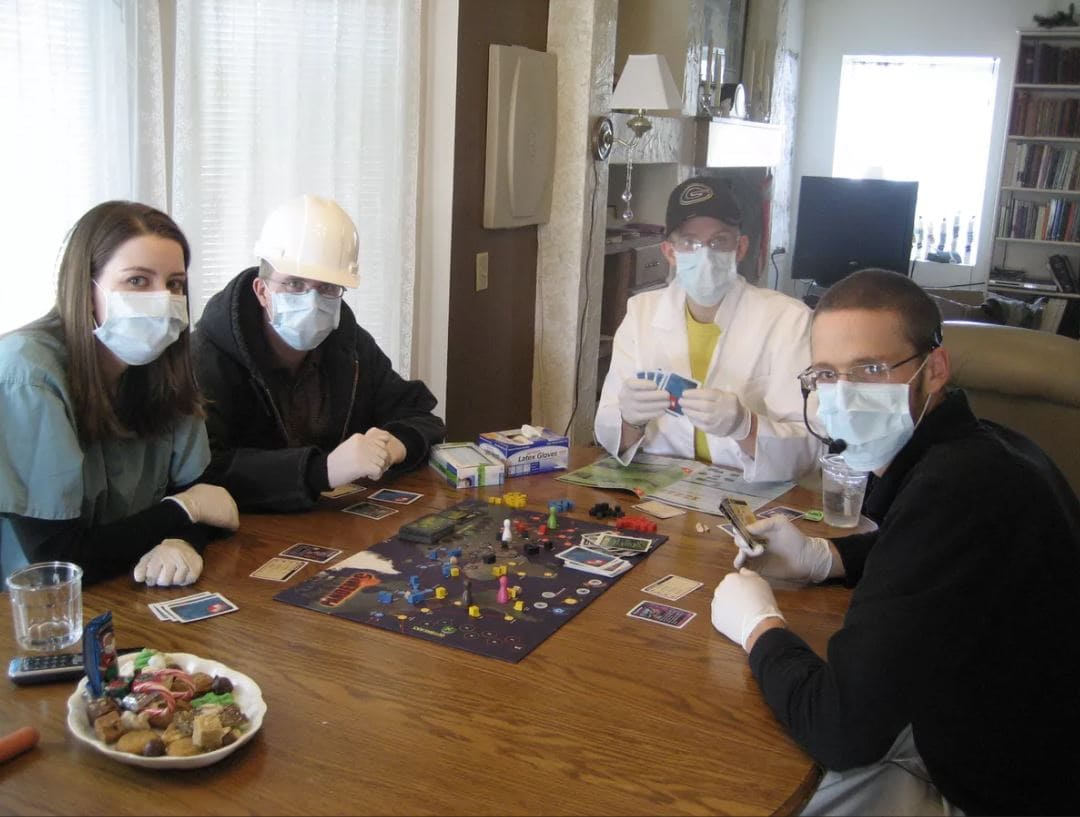 A group of four people wearing medical masks while playing Pandemic, adding a humorous touch to the game's theme.