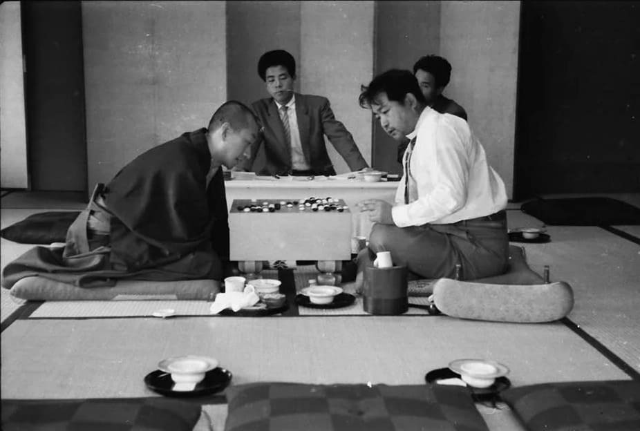 A black-and-white photo of two players engaged in a Go match, sitting on tatami mats with observers watching in the background.