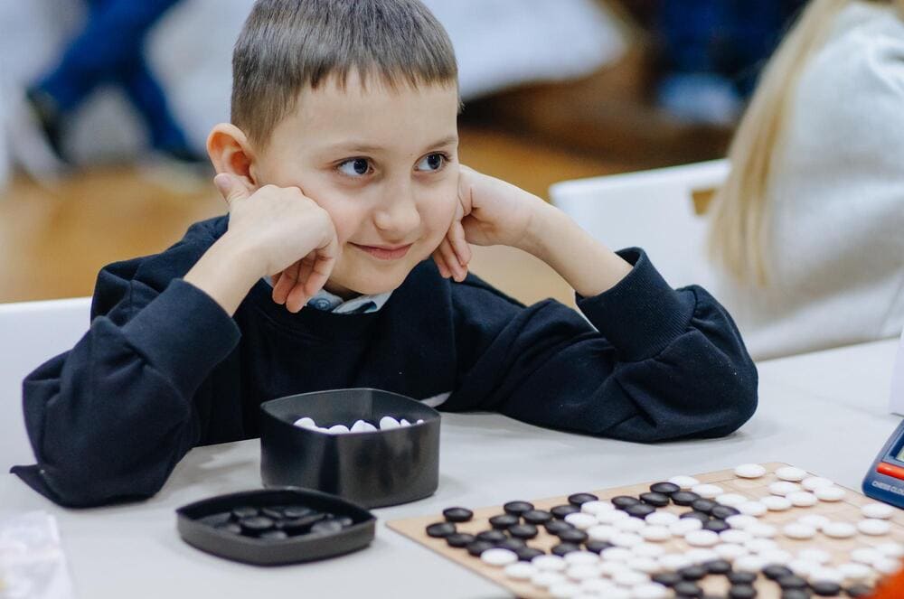 A young boy smiles while playing Go, looking at the board filled with black and white stones, enjoying the game atmosphere.