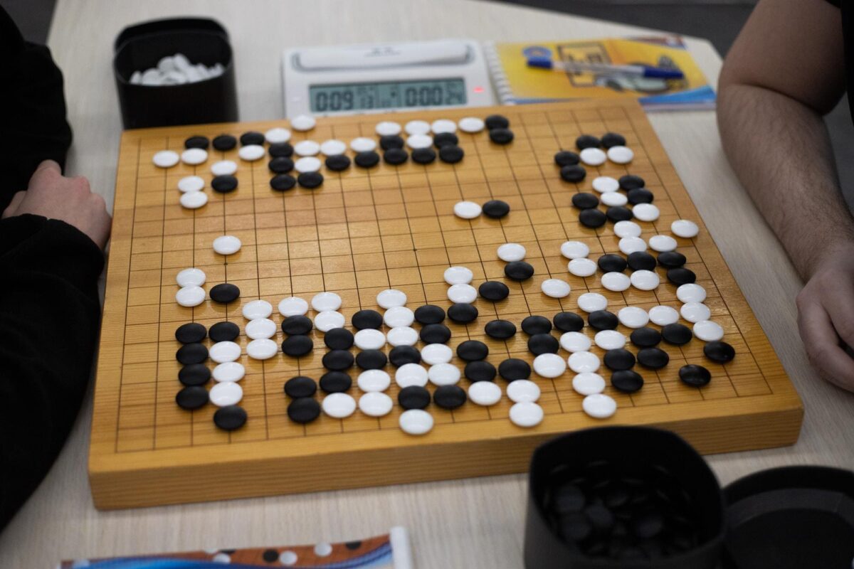 A close-up photo of a Go board with black and white stones placed across the grid, surrounded by timers and bowls of stones.