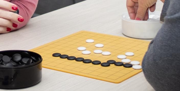 A smaller Go board with black and white stones arranged during a casual game, with hands reaching for the stones.